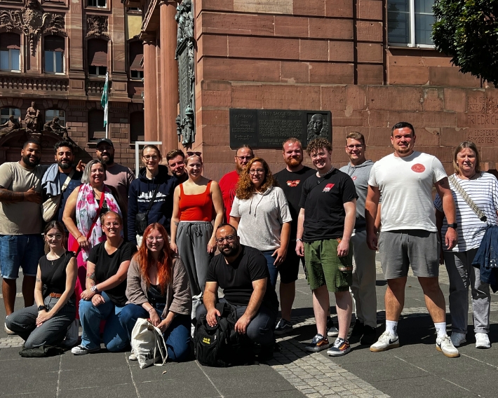 Gruppe von 20 Personen vor einem historischen Gebäude, lächelnd und in Freizeitkleidung, während eines Lehrgangs zum Ge...