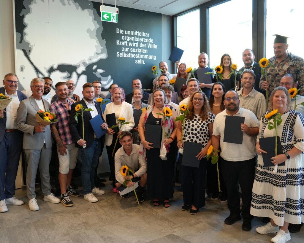Gruppenszene mit 20 Personen, die Sonnenblumen und Urkunden halten, vor einer Wand mit einem Zitat über soziale Selbstbes...