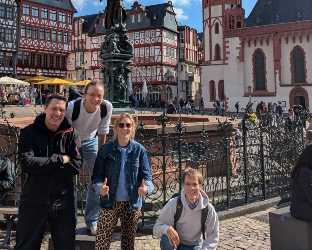 90_Ankommen in FFM - Europäische Akademie der Arbeit Vier Personen posieren vor einer historischen Brunnenanlage in Frankfurt, umgeben von Fachwerkhäusern und einer sonnigen ...