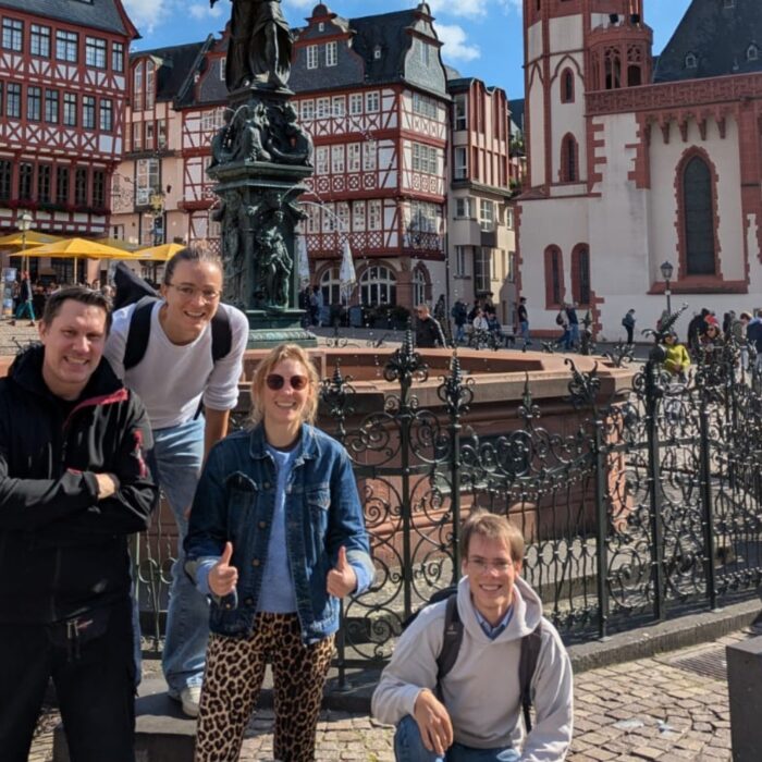 Vier Personen posieren vor einer historischen Brunnenanlage in Frankfurt, umgeben von Fachwerkhäusern und einer sonnigen ...