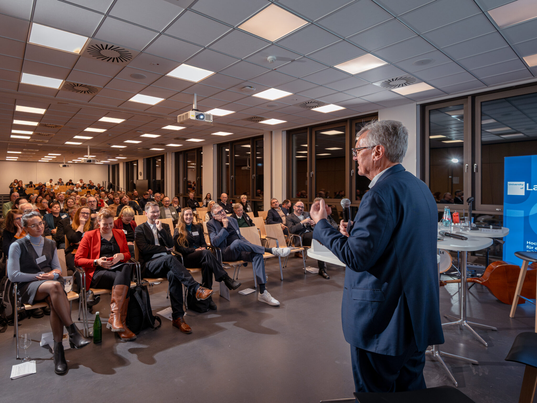 - Europäische Akademie der Arbeit Rainer Gröbel spricht vor einem Publikum in einem modernen Veranstaltungsraum, während Gäste aufmerksam zuhören.