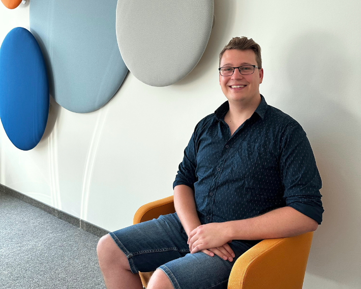 Mann mit Brille sitzt lächelnd in einem orangefarbenen Stuhl vor einer Wand mit farbigen Akustikpaneelen.